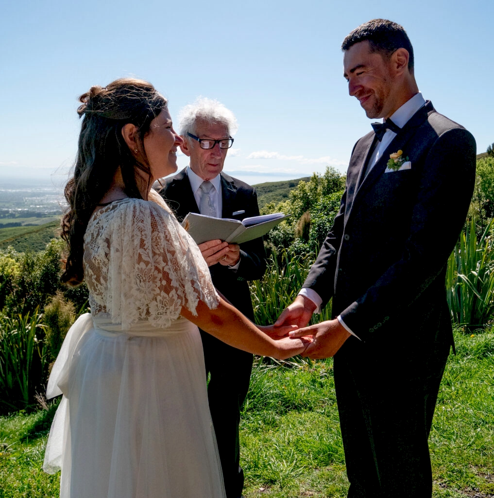 Zoe and Stewart stood in front of Sean Joyce, a Christchurch based wedding celebrant. They are stood next to the sign of the bellbird with a backdrop of the Port Hills